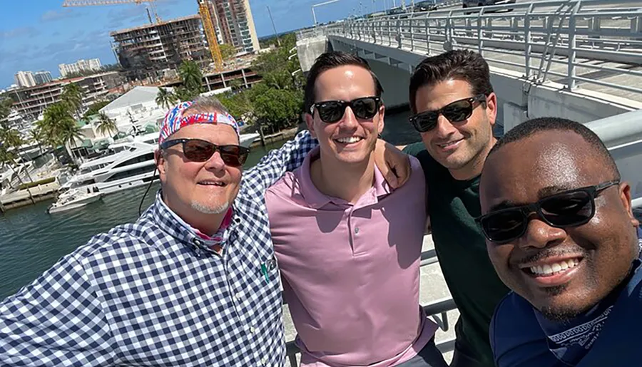 Four smiling men are taking a group selfie on a sunny day, with a marina featuring boats and construction in the background.