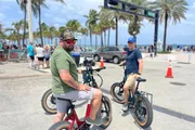 Two individuals are seen riding fat tire electric bikes on a sunny day near a palm-lined coastal area.