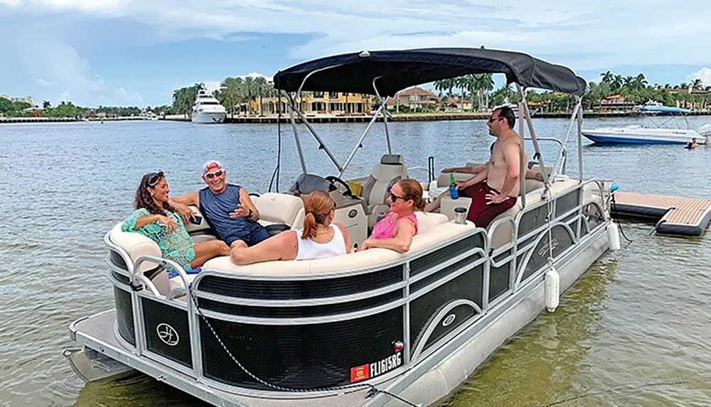 Several people are enjoying a sunny day out on a pontoon boat on calm waters with waterfront homes and another boat visible in the background