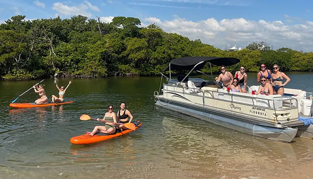 A group of people is enjoying a sunny day with activities including kayaking and relaxing on a pontoon boat near a mangrove shoreline