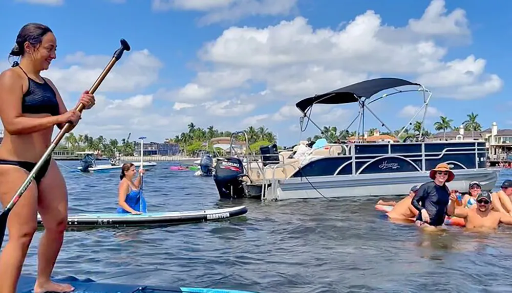 A woman is standing on a paddleboard with a paddle in hand while another is sitting on a paddleboard and a group of people are floating in the water near a moored pontoon boat under a sunny sky