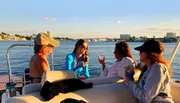 A group of friends is enjoying a boat ride and drinks on a sunny day with a scenic waterfront backdrop.