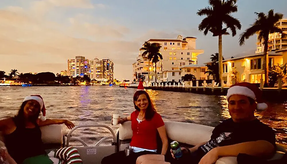 Three people are wearing Santa hats and enjoying a boat ride at dusk with a backdrop of lit buildings and palm trees