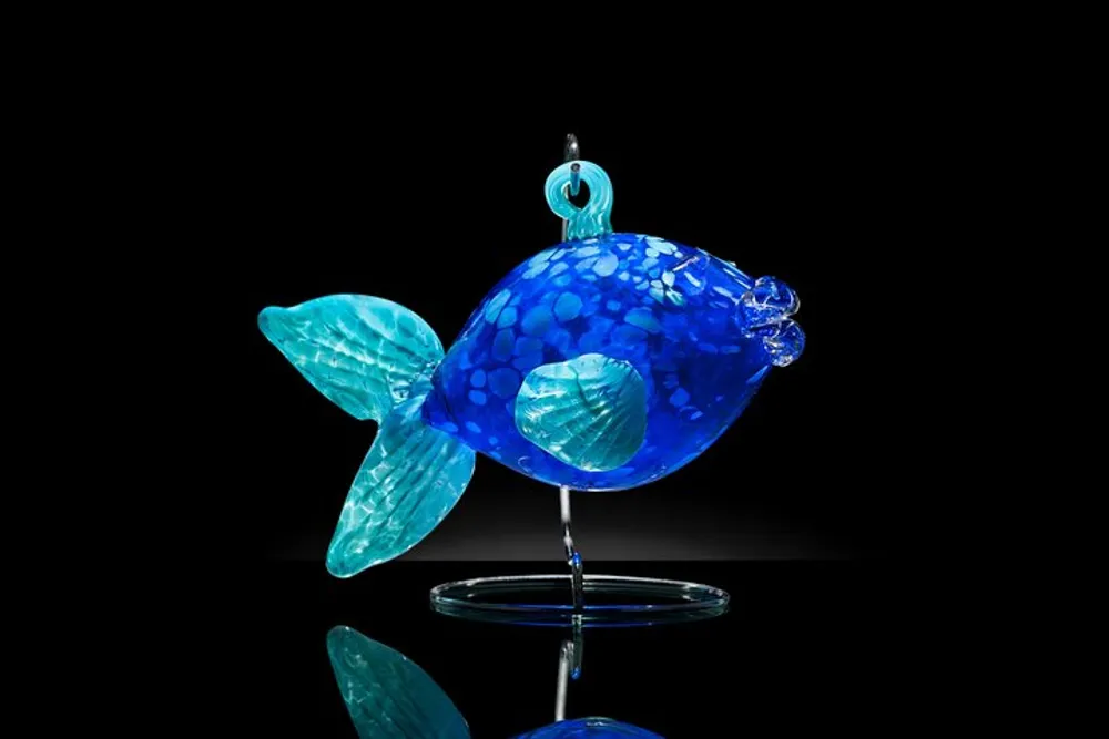 The image showcases a vibrant blue glass sculpture of a fish against a black background with a reflection on a glossy surface