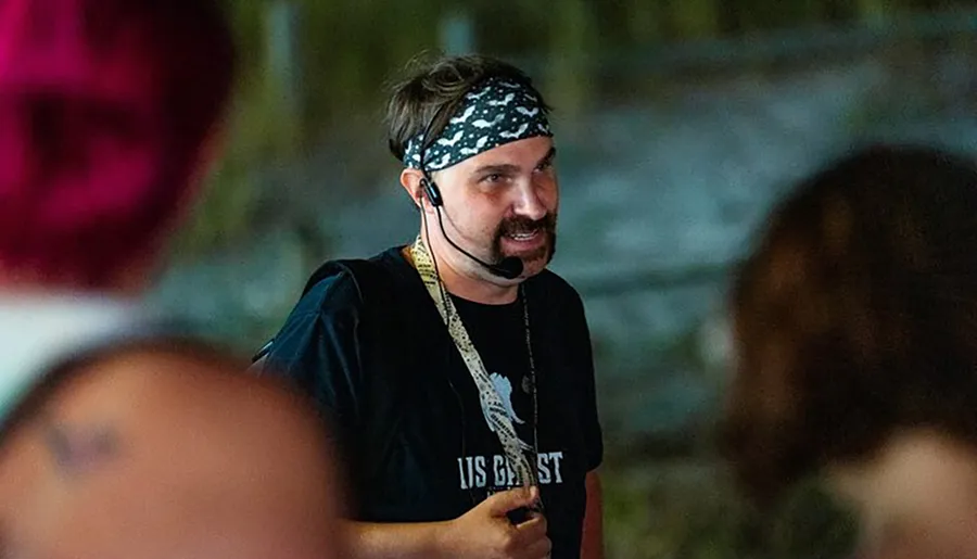 A person with a bandana and a headset microphone is speaking to an unseen audience while wearing a black T-shirt.