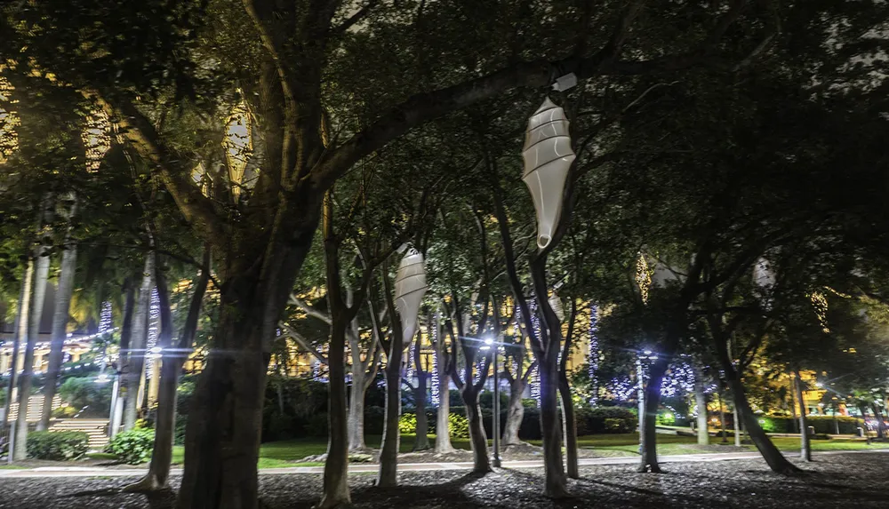 A nighttime scene in a park with large white cocoon-like installations hanging from the trees illuminated by nearby ambient lighting