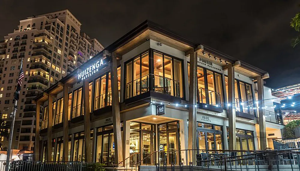 The image shows a two-story urban building at night illuminated with warm interior lights and featuring signage that reads HUIZENGA PAVILION with a high-rise building in the background