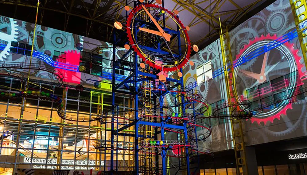 This image features a colorful kinetic sculpture with scaffolding interlaced with vivid projections of gears and mechanical elements on the walls of a modern museum interior
