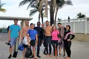 A group of smiling people in swimwear and diving gear are posing together in front of palm trees, likely about to go or having returned from a dive.