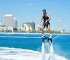 A person is using a flyboard soaring above the water with a jet of water beneath their feet against a backdrop of a sunny coastal cityscape