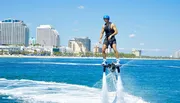 A person is using a flyboard, soaring above the water with a jet of water beneath their feet, against a backdrop of a sunny coastal cityscape.