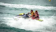 Two people are riding a jet ski across choppy water, creating a spray behind them.