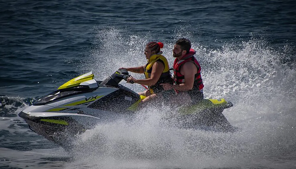 Two individuals wearing life jackets are riding a jet ski across a body of water creating a spray of water behind them