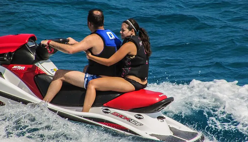 Two people are riding a red and white jet ski on a sunny day with clear blue water around them