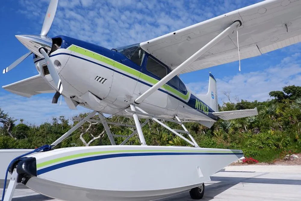 The image shows a white seaplane with green and blue stripes equipped with floats parked on a sunny day with some vegetation in the background