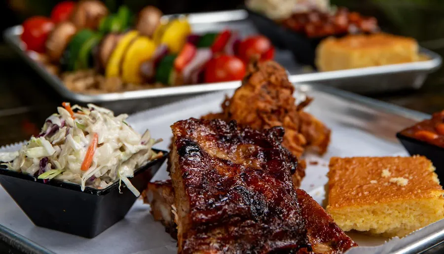 A tray with barbecued ribs, coleslaw, cornbread, and a skewer of grilled vegetables suggests a delicious BBQ meal.