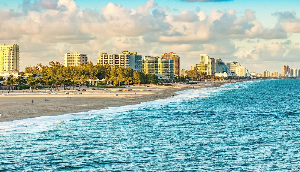 The image captures a sunny beachfront lined with high-rise buildings likely in a coastal city with the ocean in the foreground and a clear blue sky populated with a few clouds above