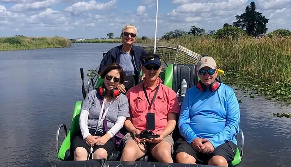 Four individuals are seated on an airboat in a waterway likely in a marsh or wetland environment with protective ear muffs on and smiling for the camera