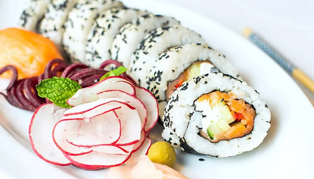 This image shows a plate of sushi rolls sprinkled with black sesame seeds accompanied by sliced radishes purple onions and a garnish with a pair of chopsticks resting beside the plate