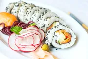 The image shows a plate of sushi rolls embellished with black sesame seeds, accompanied by pickled ginger, radish slices, wasabi, and a mint leaf, with chopsticks lying alongside.