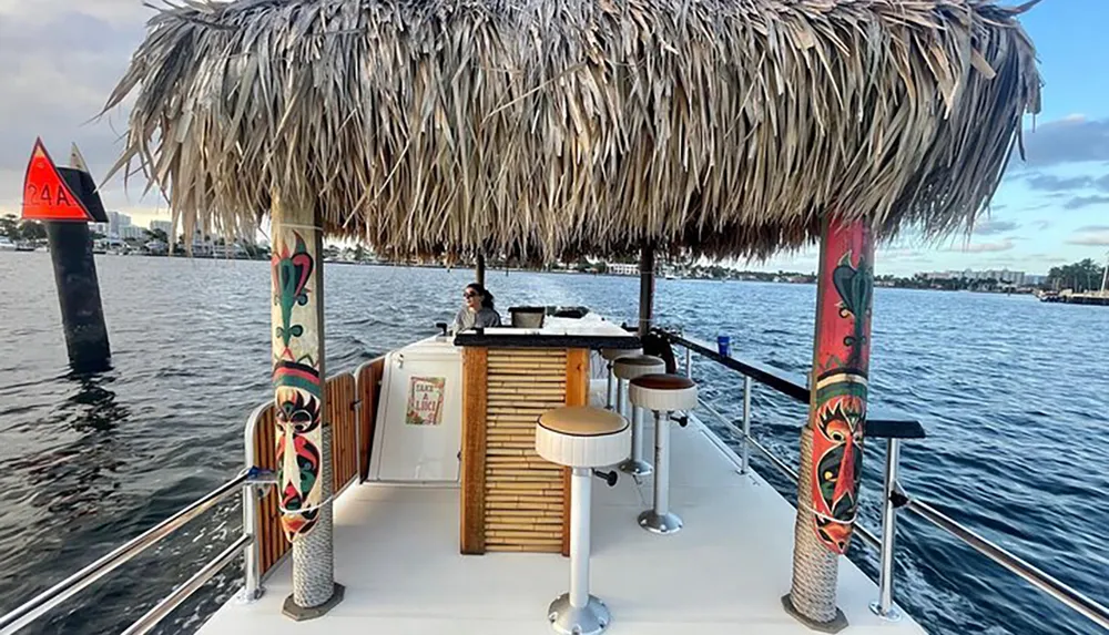 A floating tiki bar boat featuring a thatched roof and bar stools sails on the water with a person sitting at the bar and a navigational marker in the background