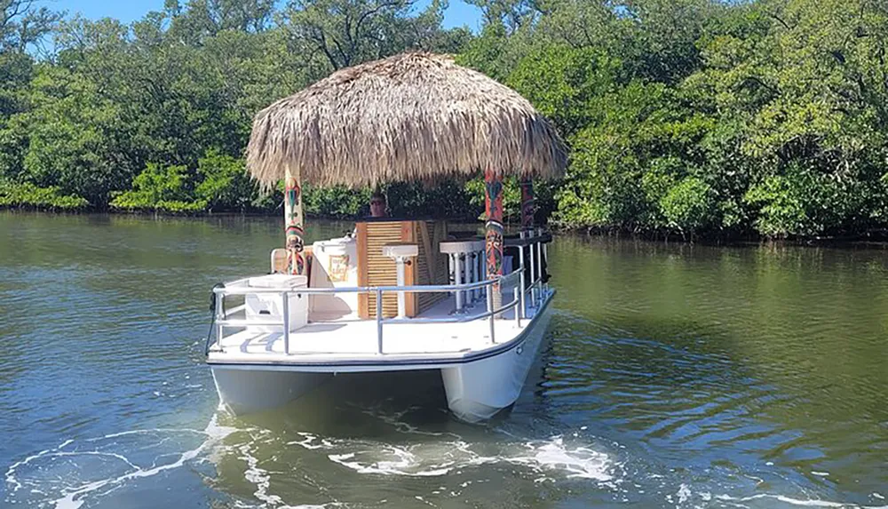 A tiki-themed pontoon boat with a thatched roof and tropical decorations is cruising on calm river waters surrounded by greenery