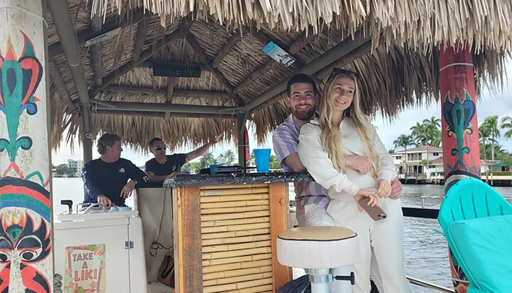 A couple is smiling and embracing near a bamboo bar with a thatch roof on a boat with two other individuals in conversation behind them