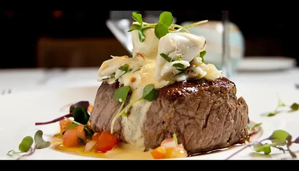 The image shows an elegantly plated steak topped with what appears to be a crab meat garnish surrounded by colorful vegetables microgreens and a sauce on a white plate