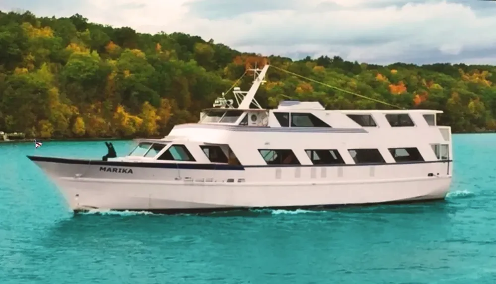 A white yacht named MARIKA is cruising on vibrant blue water with a backdrop of autumn-colored trees