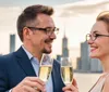 A smiling man and woman are clinking champagne glasses against a backdrop of a city skyline at sunset