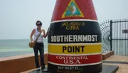 A person is standing next to the Southernmost Point buoy in Key West, Florida, indicating it's 90 miles to Cuba.