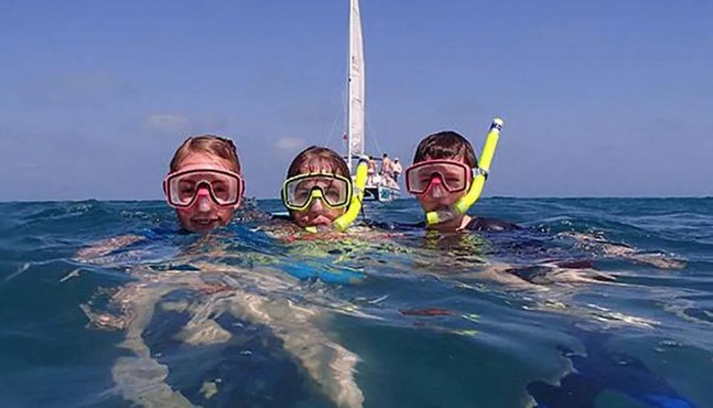 Three individuals wearing snorkeling masks and snorkels are floating in the ocean with a sailboat visible in the background