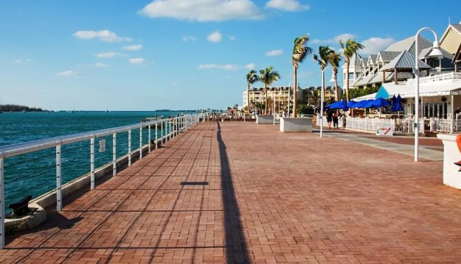 A coastal boardwalk lined with palm trees leads past outdoor seating areas and toward a serene ocean horizon under a blue sky punctuated by sparse clouds.