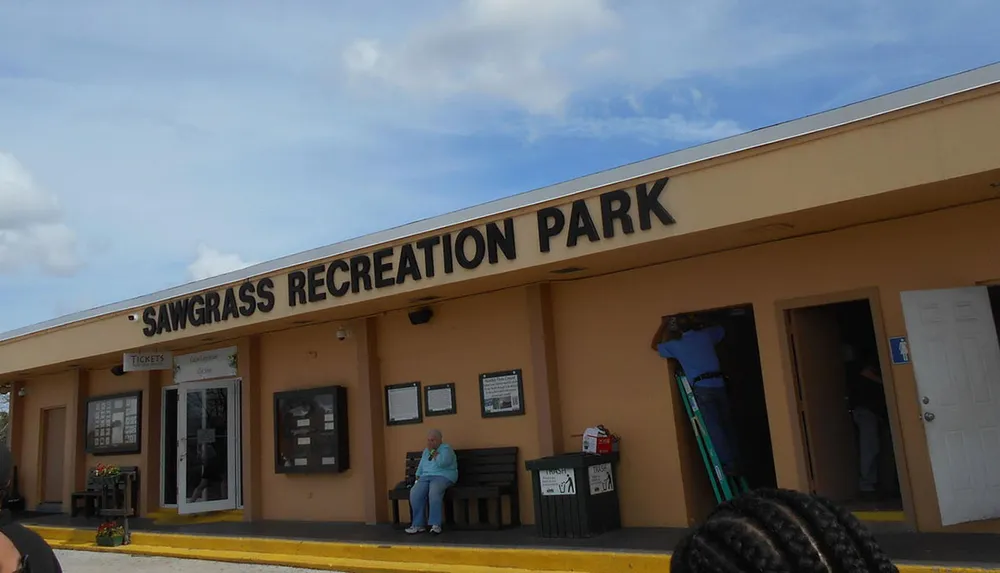 The image shows the entrance of the Sawgrass Recreation Park with a person sitting on a bench outside and another person on a ladder by the doorway