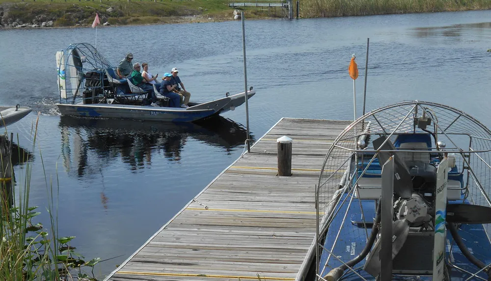 An airboat with several passengers is docked at a wooden pier on a calm body of water