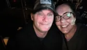 Two people are smiling for a selfie in a dimly lit environment, with one person wearing a baseball cap and the other wearing glasses.
