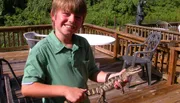 A smiling boy is holding a small alligator on a sunny deck with outdoor furniture in the background.