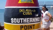 A person is posing next to the iconic Southernmost Point buoy in Key West, Florida, with the text 