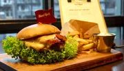 A juicy cheeseburger with bacon, lettuce, and a tomato on a wooden serving board is accompanied by a side of fries and dip against the backdrop of a casual diner setting.