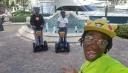 Three people are posing with two on Segways and one taking a selfie, all smiling and having a good time near a fountain and yacht.