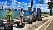 A group of people, each wearing helmets, is riding Segways in a line along a sunny, picturesque coastal sidewalk with palm trees and a city skyline in the background.