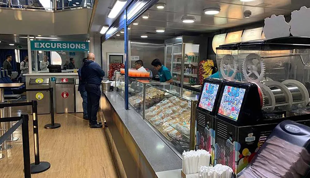 A person is standing before a counter labeled EXCURSIONS inside a ferry or cruise ship with a cafeteria setup visible and crew members in the background
