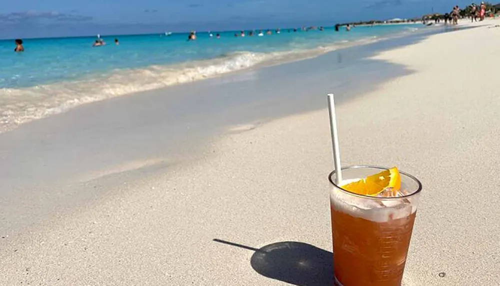 A refreshing beverage with a slice of orange and a straw is placed on a sandy beach with people and the turquoise sea in the background