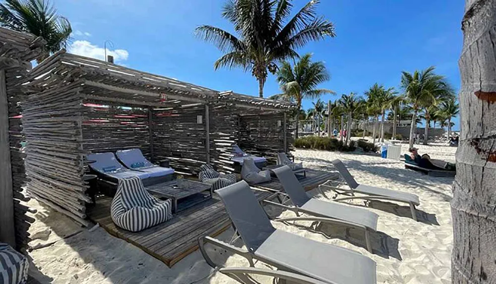 The image shows a relaxed beach setting with a rustic wooden cabana and sun loungers under the shade of palm trees suggesting a leisurely vacation spot
