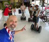 A person is taking a selfie with a group of people all wearing helmets and standing on Segways in a rental shop with a joyful and excited expression