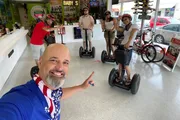 A person is taking a selfie with a group of people, all wearing helmets and standing on Segways, in a rental shop with a joyful and excited expression.