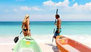 Two people stand by clear kayaks on a sandy beach, ready to paddle out into the serene turquoise waters.