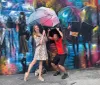 Two people are playfully interacting with a colorful street mural that depicts an urban scene with pedestrians holding umbrellas