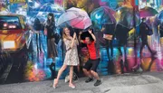 Two people are playfully interacting with a colorful street mural that depicts an urban scene with pedestrians holding umbrellas.
