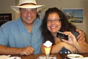 A smiling duo, one wearing a straw hat, sits at a table with a dessert in front of them, as one holds up a smartphone displaying an image.
